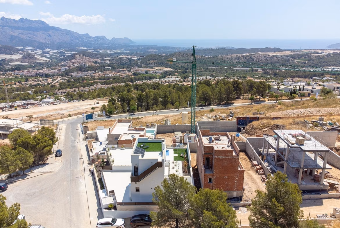 Stunning-New-Build-Villas-in-Polop-with-Sea-Views-1 New build villa under construction in Polop with flat rooftops, nearby buildings, trees, and distant sea views.