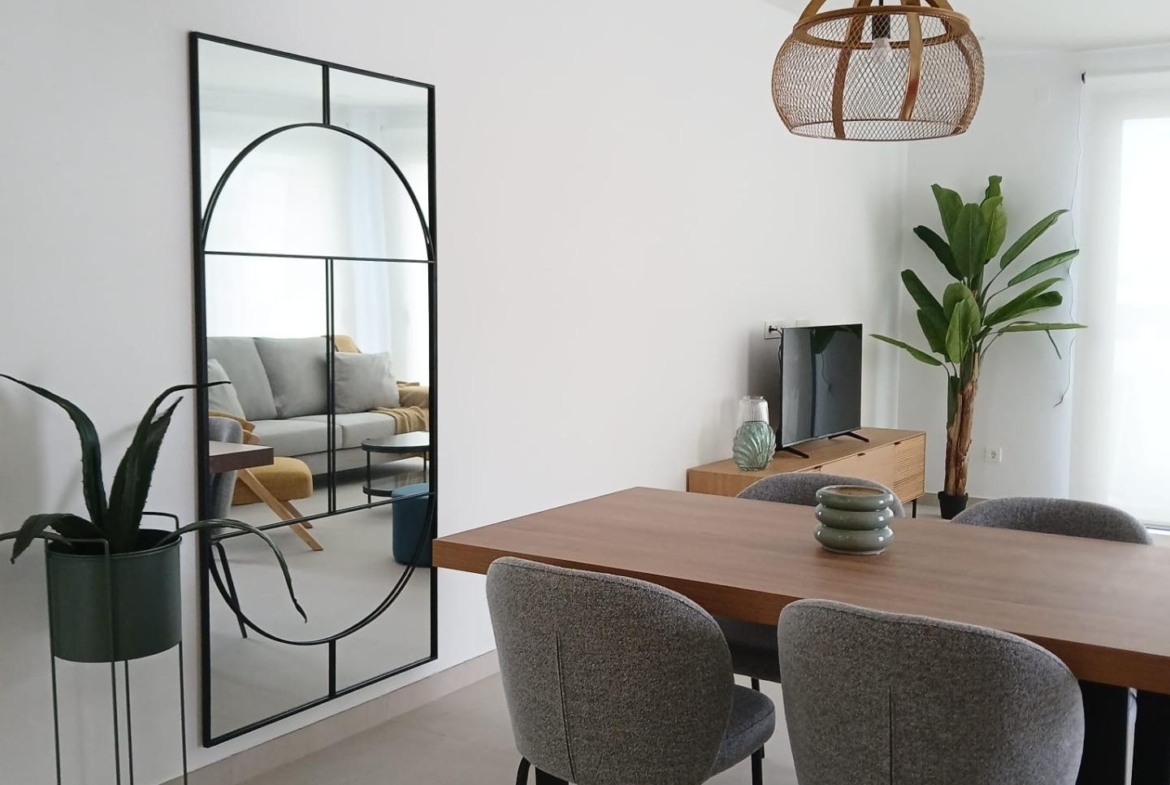 Modern dining area with wooden table, four gray chairs, decorative plants, and large mirror in Bigastro quad villa interior
