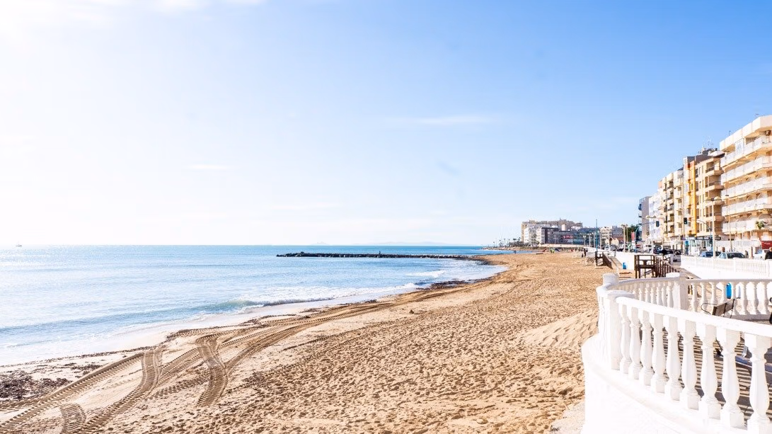 NEW-BUILD-RESIDENCIAL-IN-LOS-BALCONES-TORREVIEJA-17 Sandy beach and calm sea view from a top floor apartment in Los Balcones, Torrevieja with nearby buildings and clear sky.