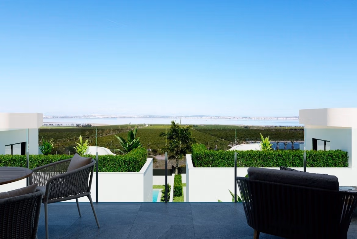 NEW-BUILD-RESIDENCIAL-IN-LOS-BALCONES-TORREVIEJA-6 Terrace with seating and glass railing overlooking green fields and water from a top floor apartment in Los Balcones, Torrevieja.
