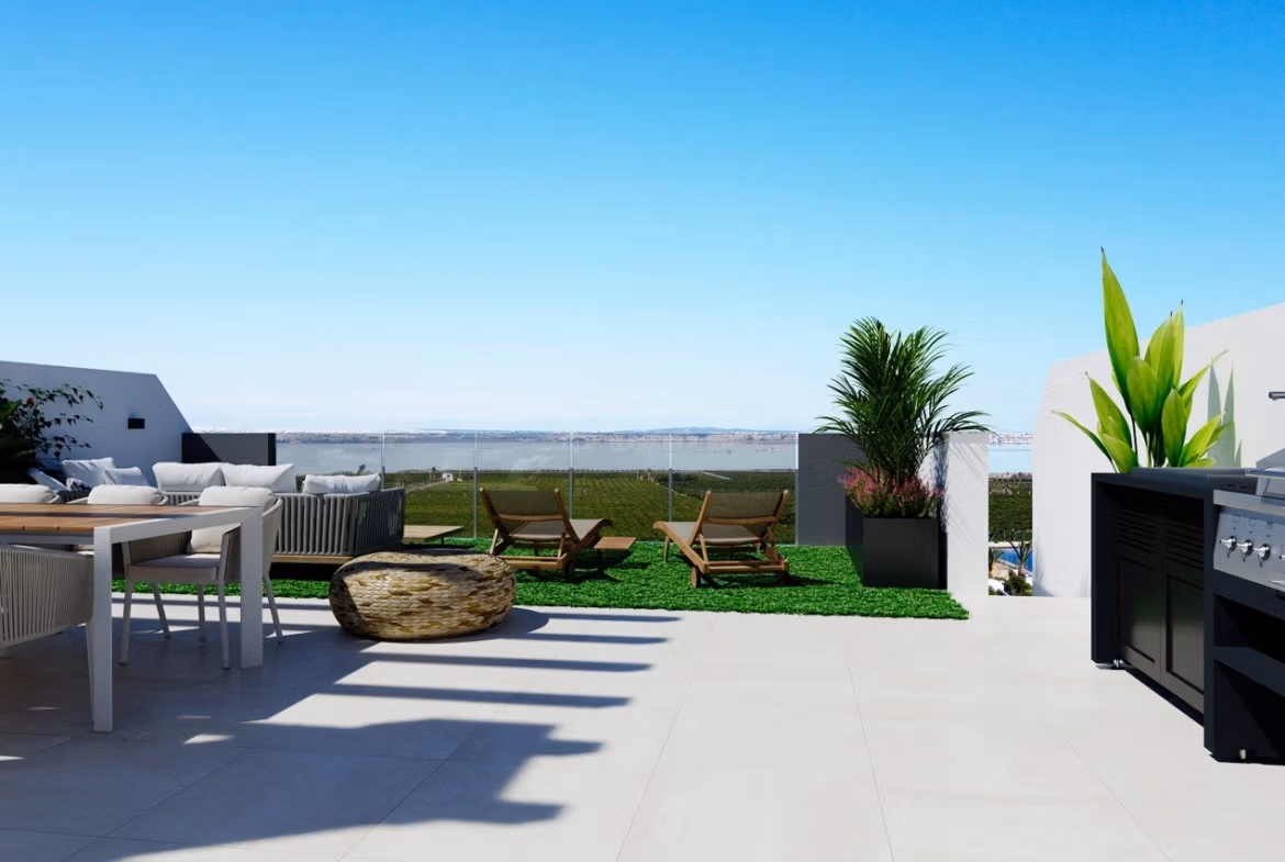 NEW-BUILD-RESIDENCIAL-IN-LOS-BALCONES-TORREVIEJA-7 Top floor apartment solarium in Los Balcones, Torrevieja with outdoor seating, sun loungers, plants, and distant sea view.