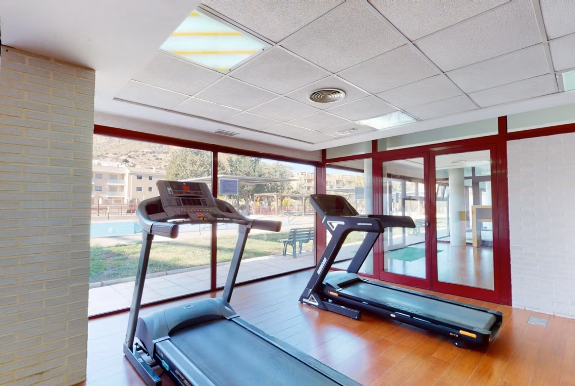 Bright, modern gym with panoramic windows overlooking the landscaped pool area in a Key Ready 1 Bedroom Apartment, Villanueva del Rio Segura.