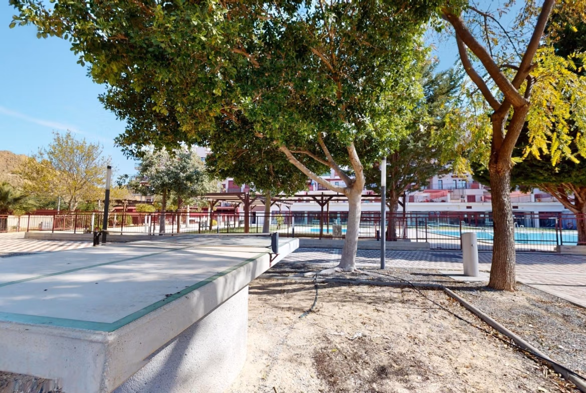 Bright outdoor recreation area with shaded seating and mature trees, part of a charming 1-bedroom apartment complex in Villanueva del Rio Segura near Archena