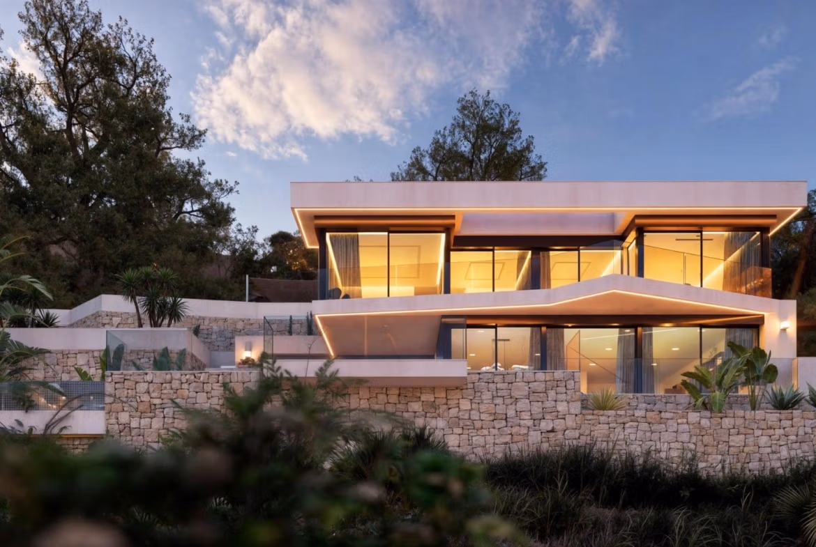 Stunning modern 4-bedroom villa in Benissa Costa with expansive glass walls, warm interior lighting, and landscaped stone terraces.
