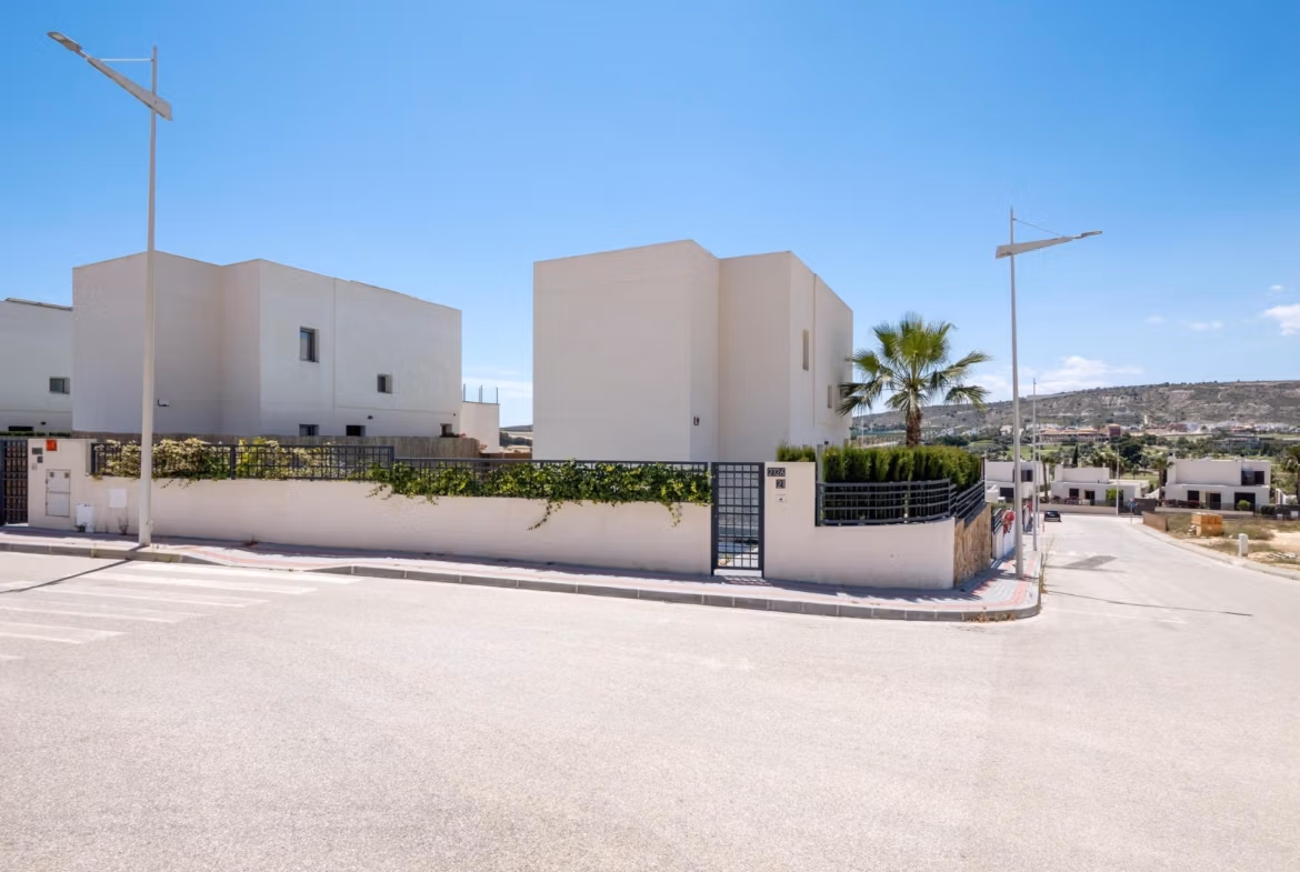 Modern white 4-bedroom villa with gated entrance and landscaped corner plot in sunny Algorfa, La Finca, with clear blue skies.