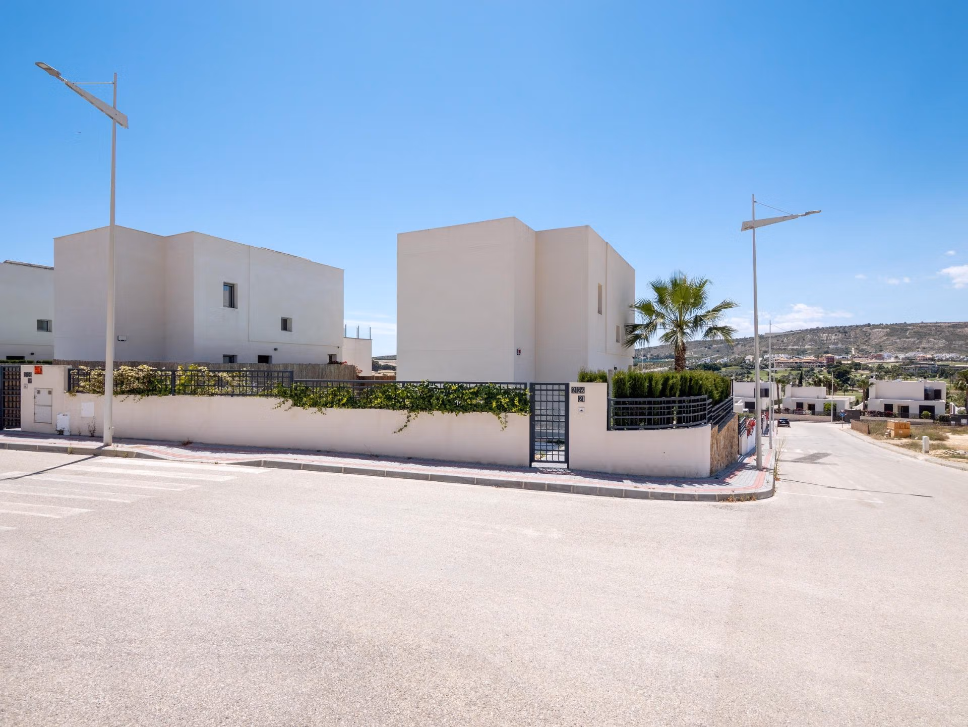Modern white 4-bedroom villa with gated entrance and landscaped corner plot in sunny Algorfa, La Finca, with clear blue skies.