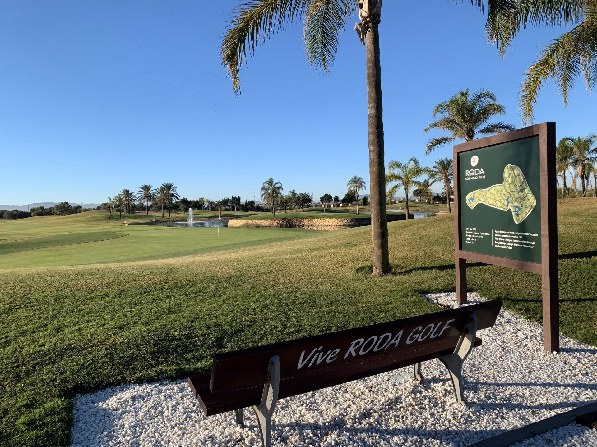 Sunlit golf course overview with lush greens and palm trees near a bench, highlighting the serene setting of Roda Golf, San Javier bungalow.