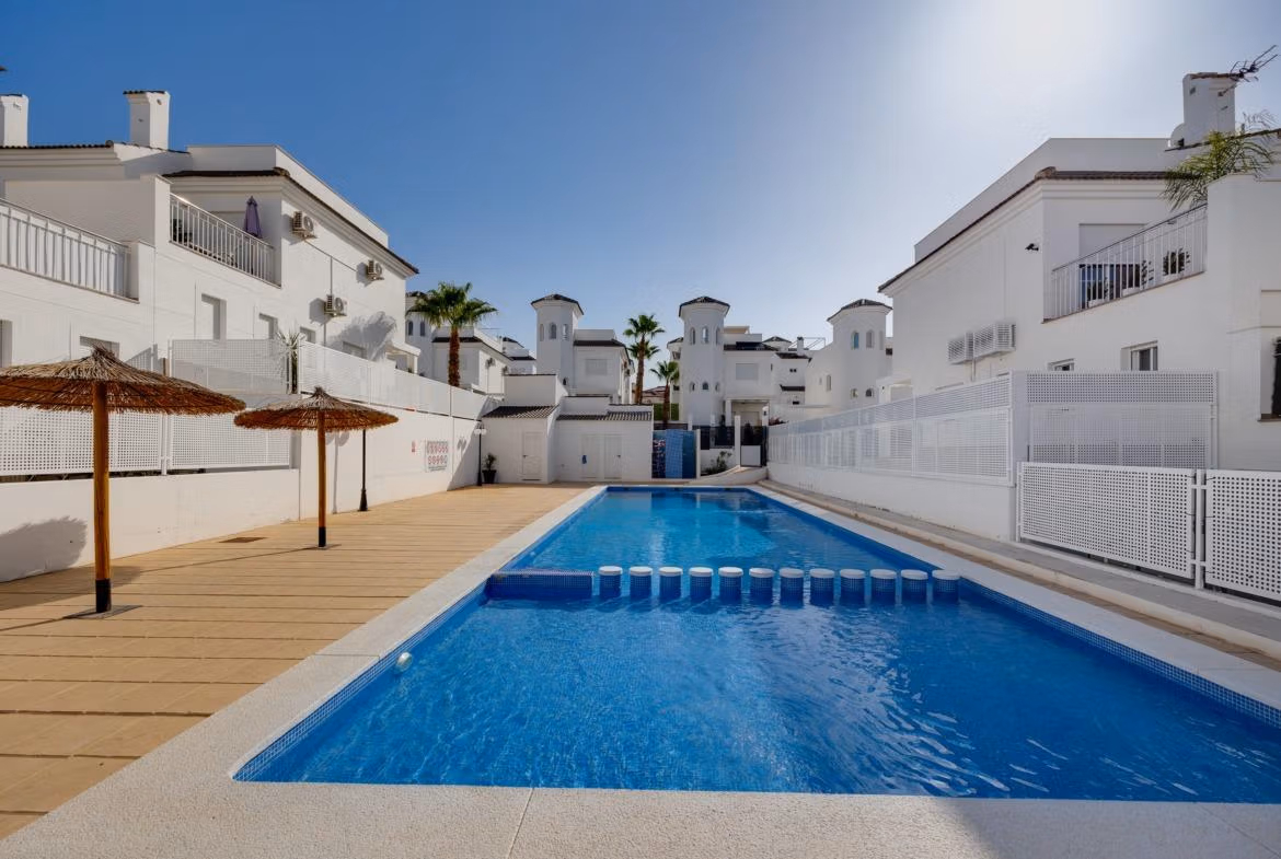 Sun-drenched communal pool with shaded loungers surrounded by elegant white 3-bedroom villas in El Oasis, San Fulgencio