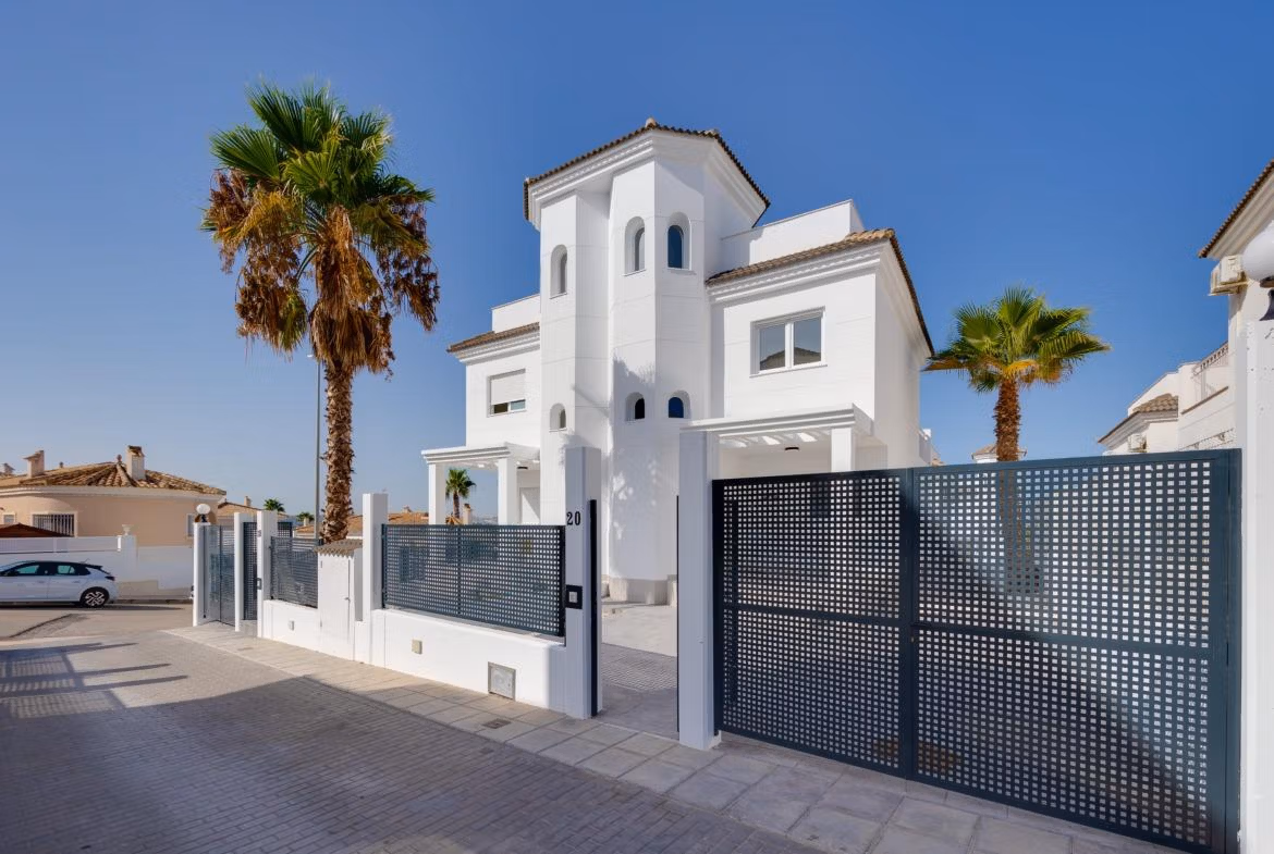 NEW-BUILD-RESIDENTIAL-IN-SAN-FULGENCIO-2 Modern, elegant 2-bedroom semi-detached villa with white facade, secure gated entrance, and palm trees in El Oasis La Marina Urbanisation