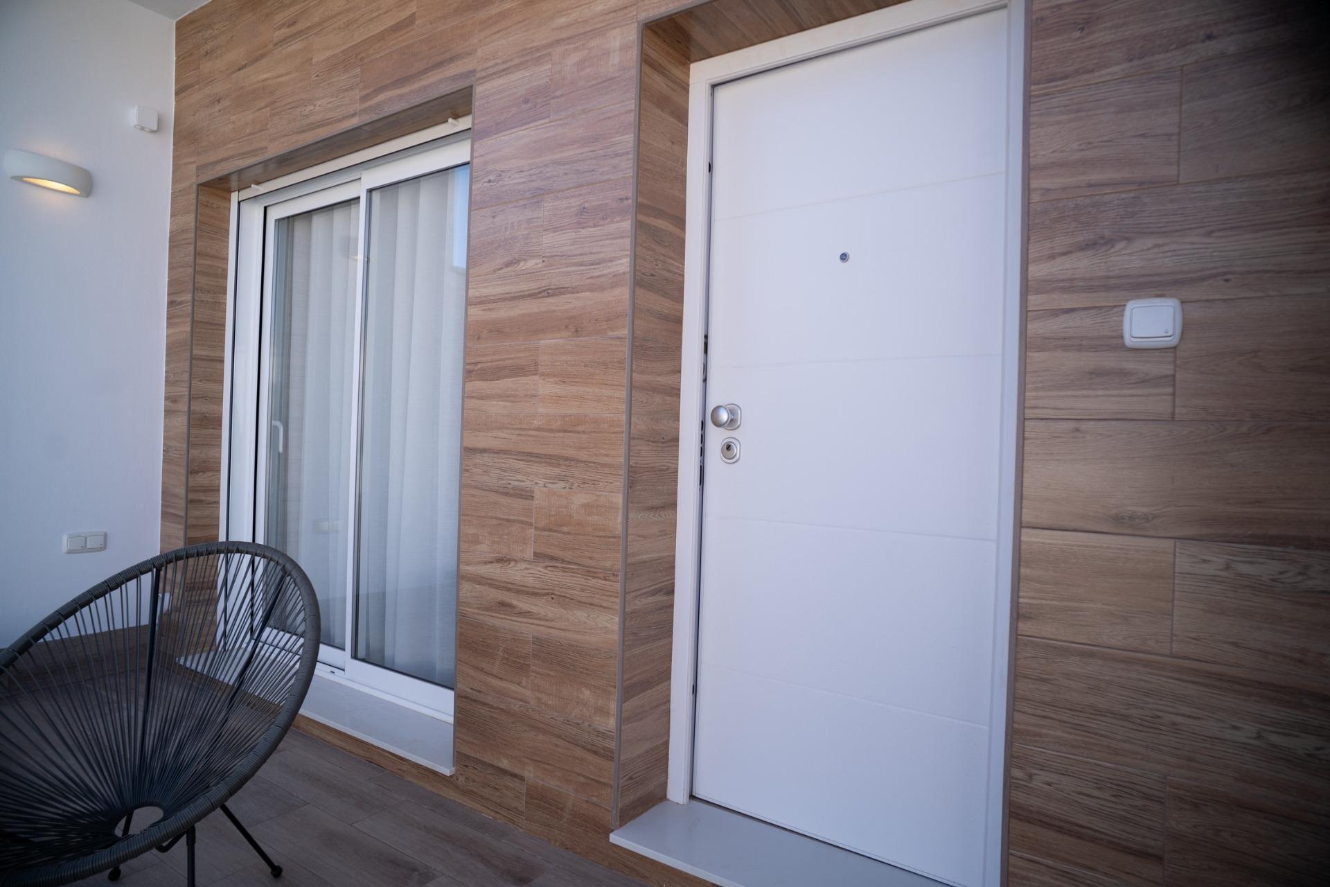 NEW-BUILD-TOWNHOUSES-IN-SAN-JAVIER-9 Modern sun-drenched terrace with sleek white door, wood-effect cladding, and stylish black chair in San Javier townhouses.