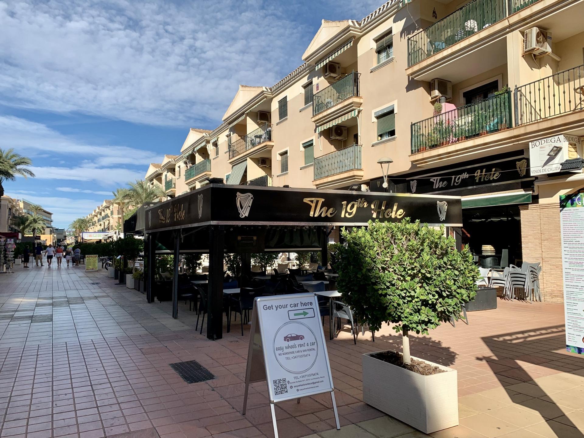 Vibrant sunlit street with elegant cafes and balconies in Los Alcázares, near exclusive new build villas with private pools.