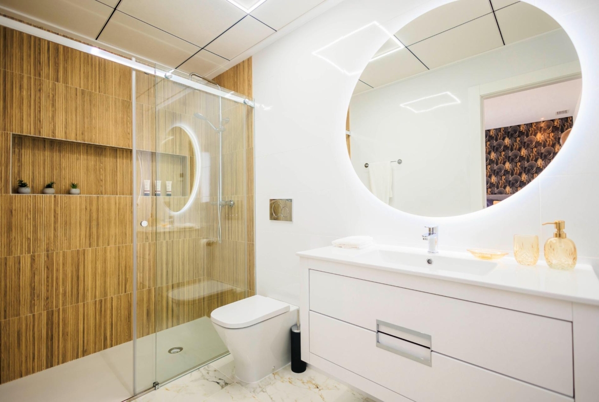 NEW-BUILD-VILLAS-IN-PILAR-DE-LA-HORADADA-ON-THE-GOLF-COURSE-20 Modern bathroom with illuminated round mirror and elegant wood-paneled shower in new build villas in Pilar de la Horadada golf course