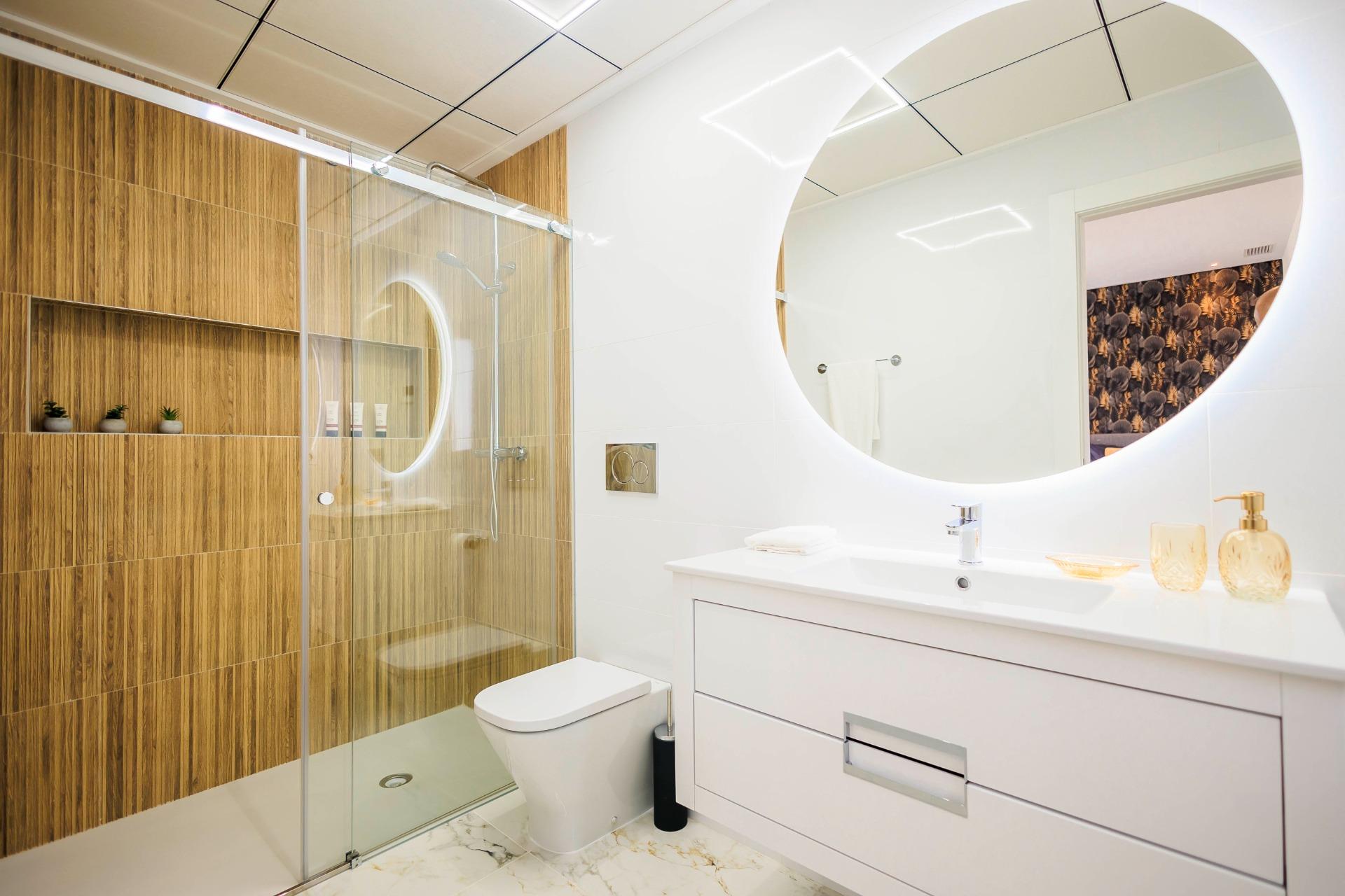 NEW-BUILD-VILLAS-IN-PILAR-DE-LA-HORADADA-ON-THE-GOLF-COURSE-20 Modern bathroom with illuminated round mirror and elegant wood-paneled shower in new build villas in Pilar de la Horadada golf course