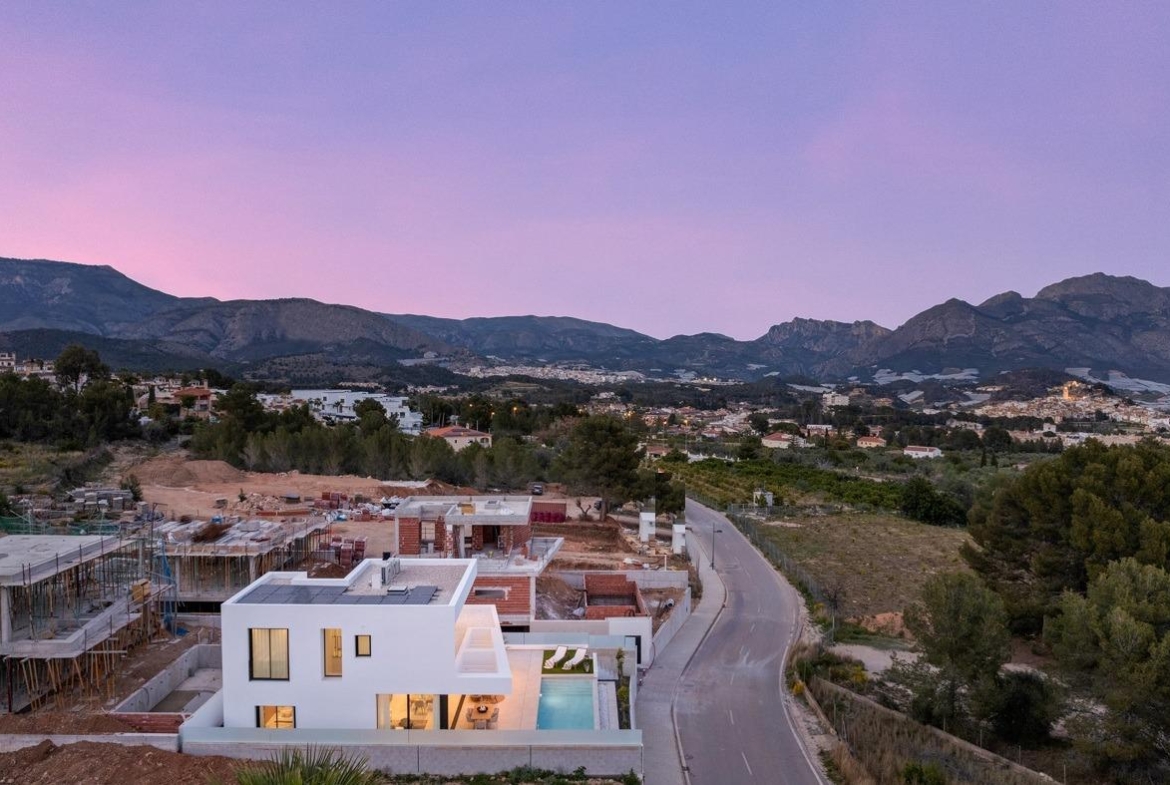 Modern white 3-bedroom villa with private pool, nestled in landscaped surroundings by serene road in Polop, Costa Blanca at sunset.