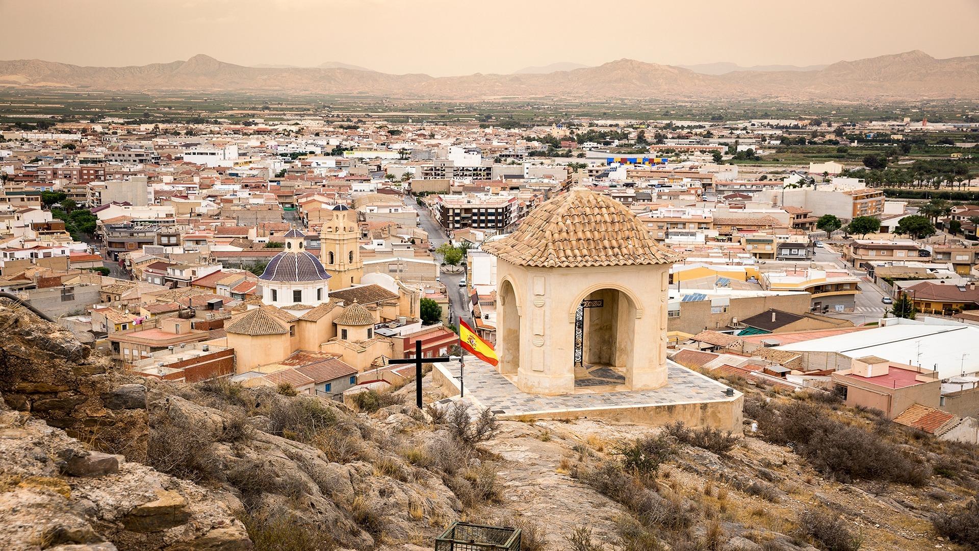 Panoramic view over historic Cox, Alicante with traditional rooftops and distant mountains under a soft, warm sky