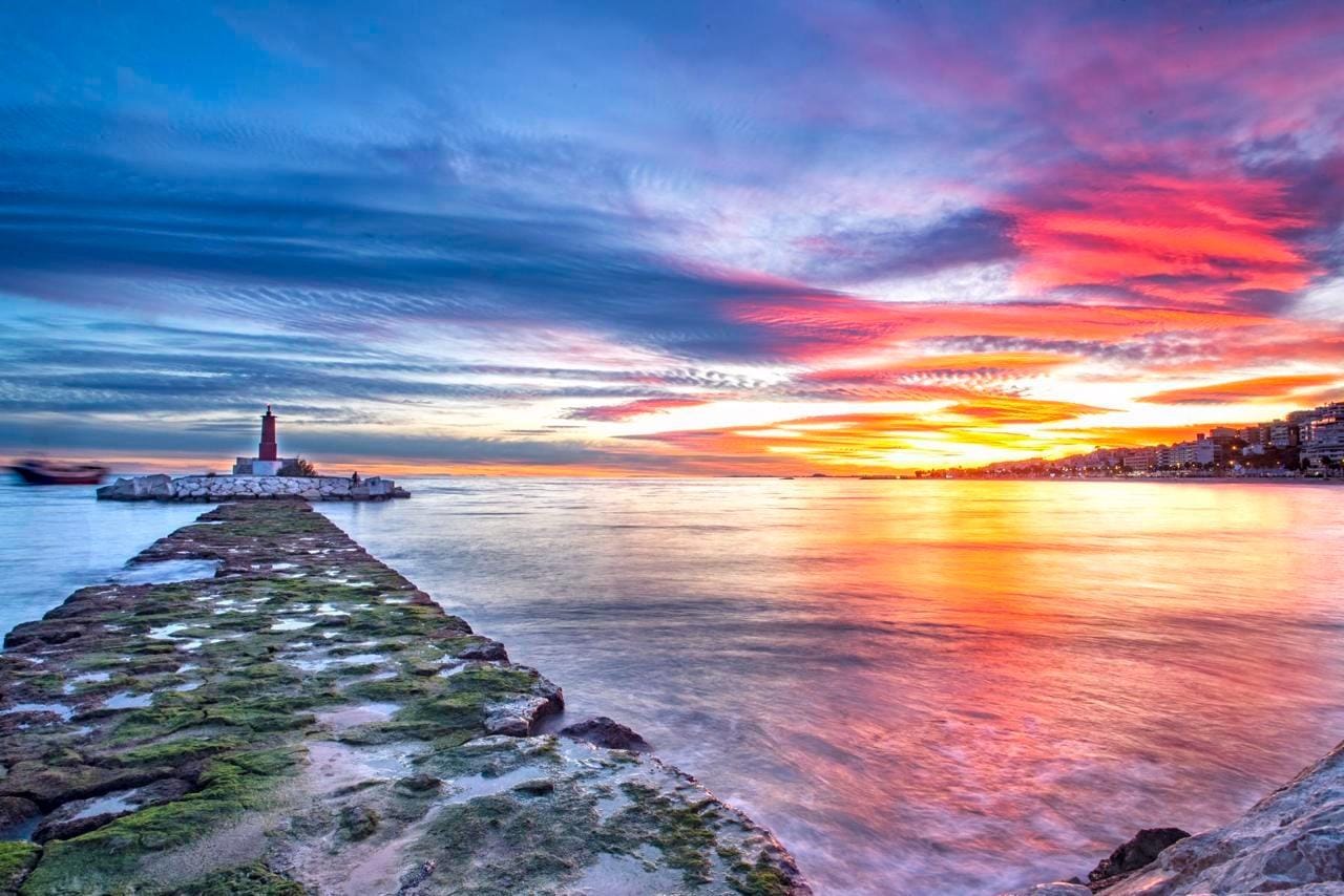 Sunset over the sea with a stone breakwater leading to a lighthouse