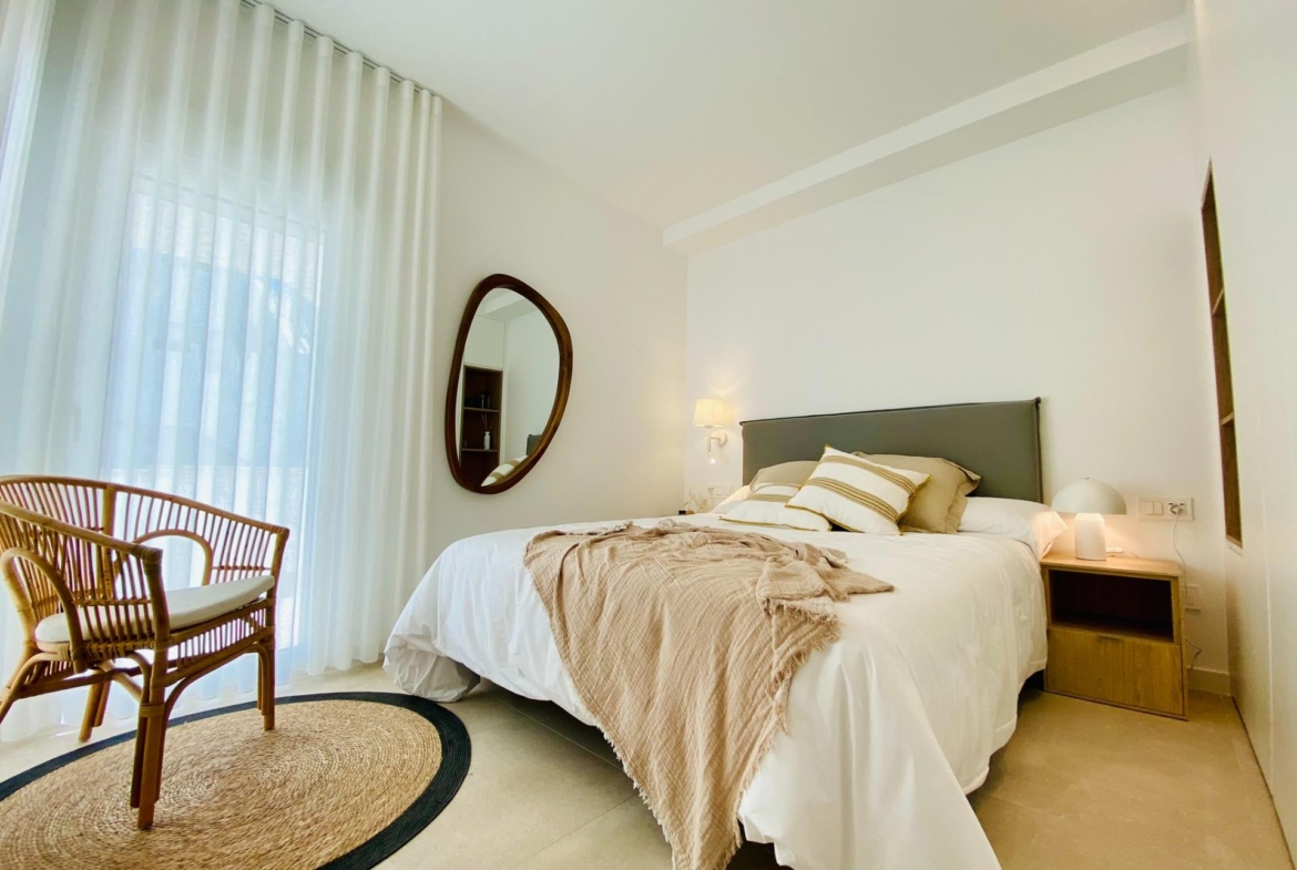 Bright bedroom with double bed, bedside table, round wall mirror and wicker chair by window